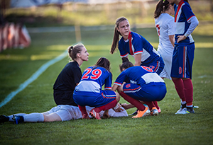 Group of female soccer players and referee gathering around injured player on playing field.