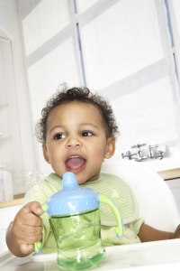 Niño que comienza a caminar, en una silla alta, tratando de alcanzar una taza con tapa.